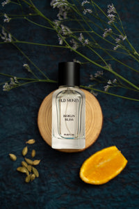 Perfume bottle labeled 'Old Money Berlin Bliss' on a dark surface with flowers and an orange slice.